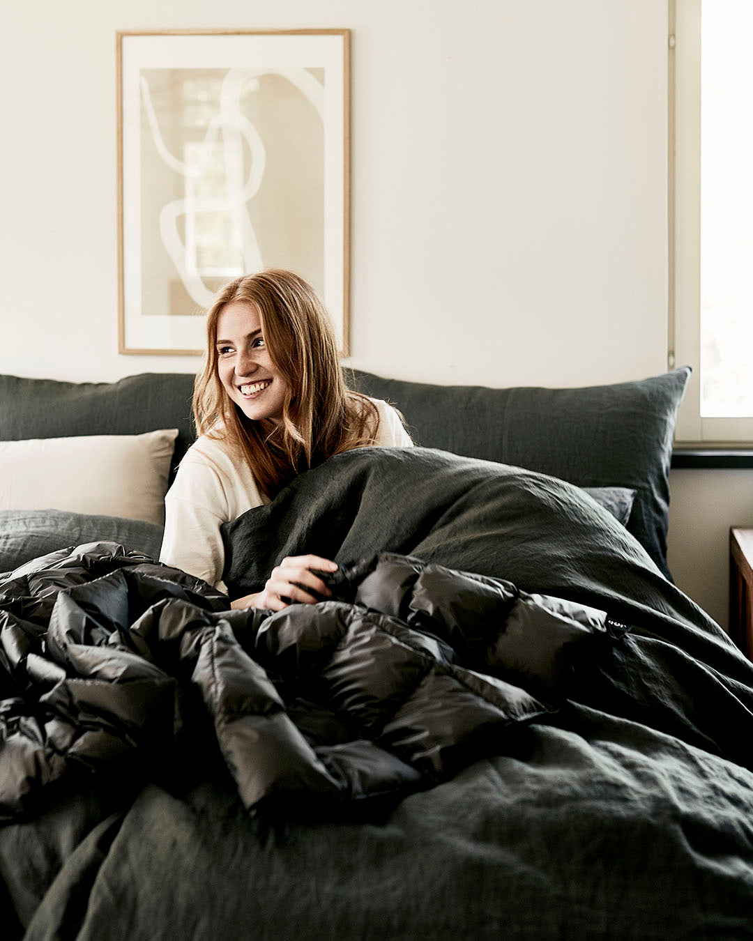 Hymyilevä henkilö istuu sängyllä tummanharmaiden vuodevaatteiden keskellä ja on kääriytynyt kiiltäväpintaiseen ruututikattuun Kulkuri-untuvatorkkupeittoon (väri: antrasiitti). Taustalla vaalea seinä, graafinen taulu ja ikkuna, josta tulee luonnonvaloa huoneeseen.