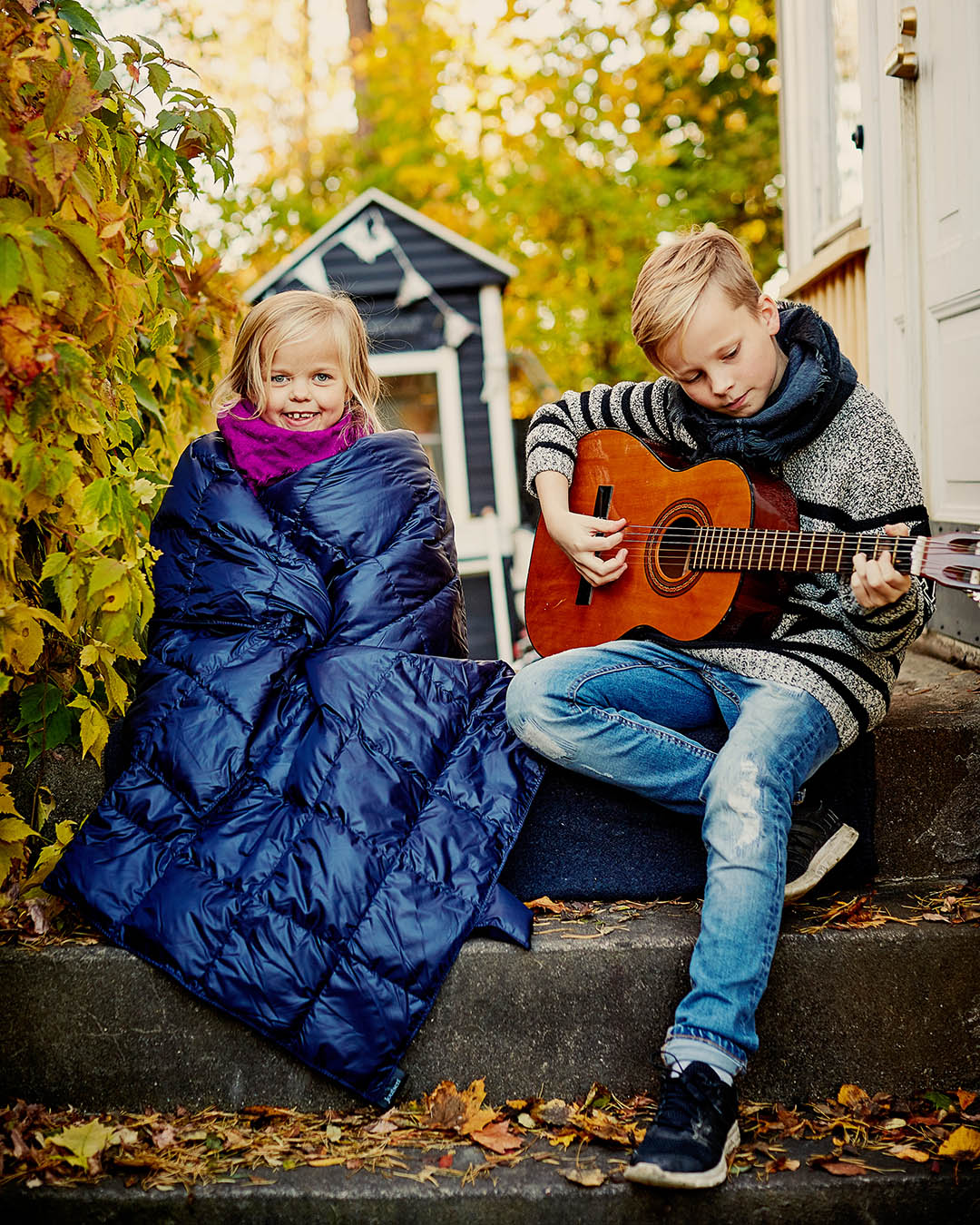 Kaksi lasta istuu rappusilla. Toinen lapsi on kääriytynyt tummansiniseen Kulkuri-untuvatorkkupeittoon ja hymyilee kameraan, toisella on harmaa villapaita ja huivi, ja hän soittaa kitaraa. Taustalla näkyy värikkäitä syyspensaita ja pieni mökki.