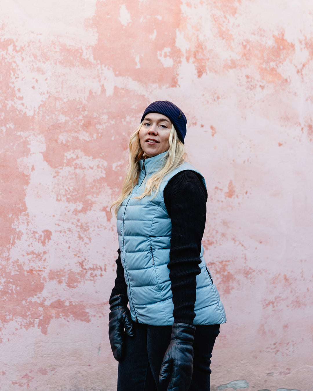 Nainen seisoo vaaleanpunaisen, seinän edessä pukeutuneena kissankellonsiniseen Lady-untuvaliiviin. Liivissä on kevyesti kiiltävä pinta ja vaakasuuntaiset tikkaukset. Asuun kuuluu myös musta pipo, neule, housut ja nahkahanskat.