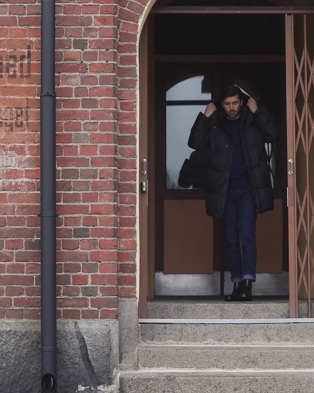 Mies kulkee kaupunkimaisessa maisemassa yllään musta Koski-untuvatakki. Takin vetoketju on auki ja alta näkyy tumma neule ja vaalean kauluspaidan kaulukset. Mies on pukeutunut tummansinisiin farkkuihin ja nahkaisiin nilkkureihin.