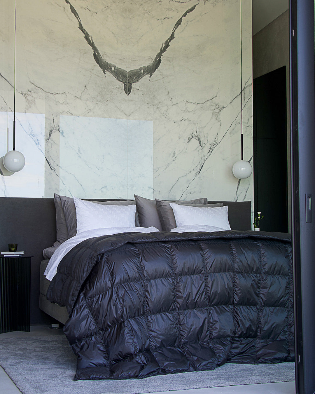 Sängyllä on tikattu Henkäys-päiväpeitto, joka ulottuu reunoilta alas lattialle. Peitto on tumman harmaa (väri: antrasiitti), ja sen pinta heijastaa kevyesti valoa. Taustalla on näyttävä marmorikuvioinen seinä, ja sängyllä on useita harmaita ja valkoisia tyynyjä.