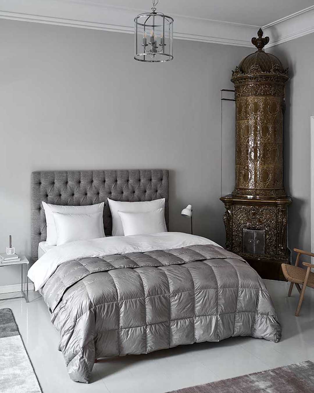 Sängyllä on tikattu Henkäys-päiväpeitto, joka laskeutuu pehmeästi reunoilta kohti lattiaa. Peitto on harmaa ja ruututikattu. Taustalla näkyy harmaa, napitettu sängynpääty, valkoisia tyynyjä ja vanha koristeellinen kaakeliuuni.