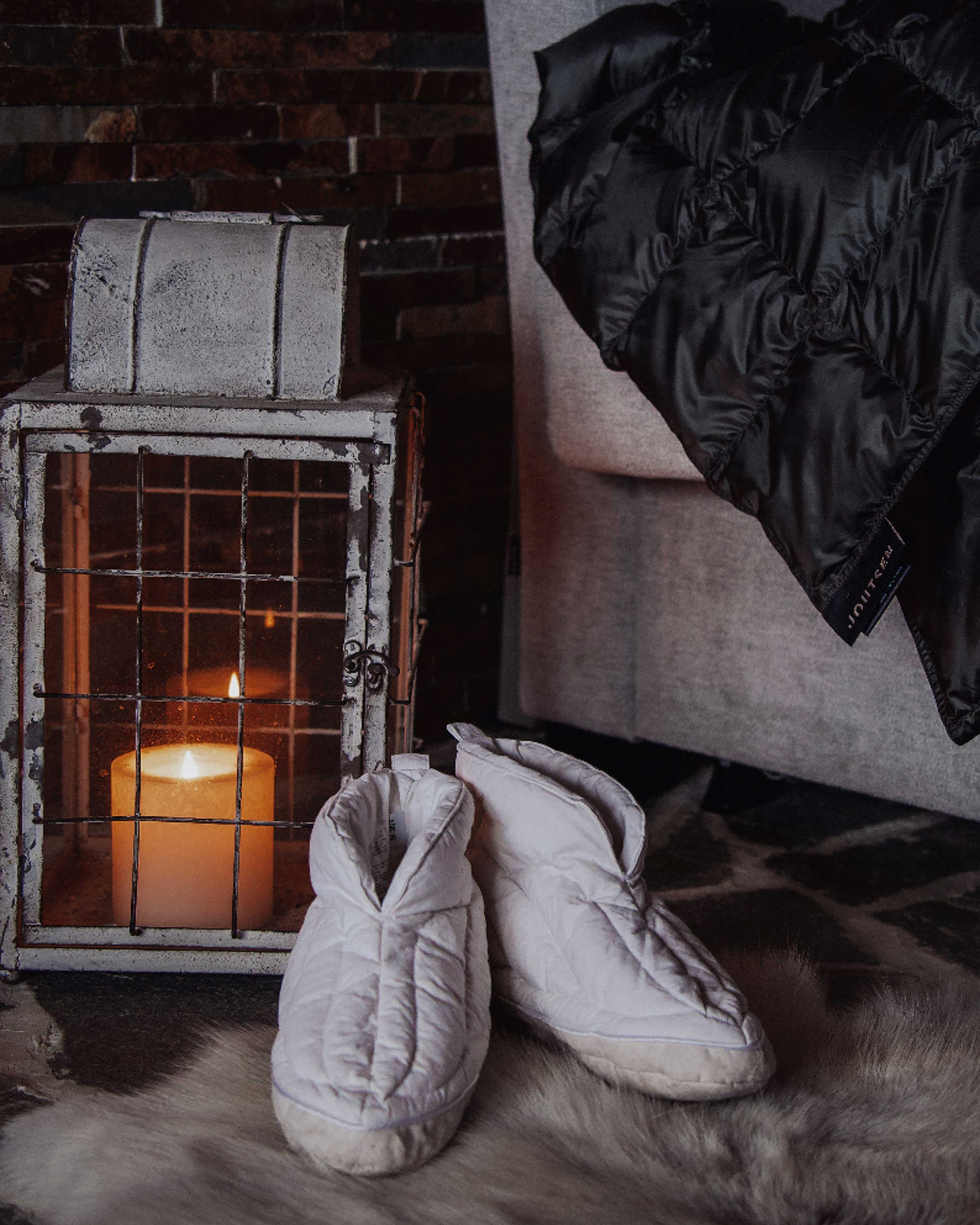 Valkoiset Kuiske-untuvatossut lepäävät taljan päällä takan edessä, jossa palaa kynttilä vanhan metallilyhdyn sisällä. Vieressä näkyy tumma Kulkuri-torkkupeitto nojatuolin käsinojalla.