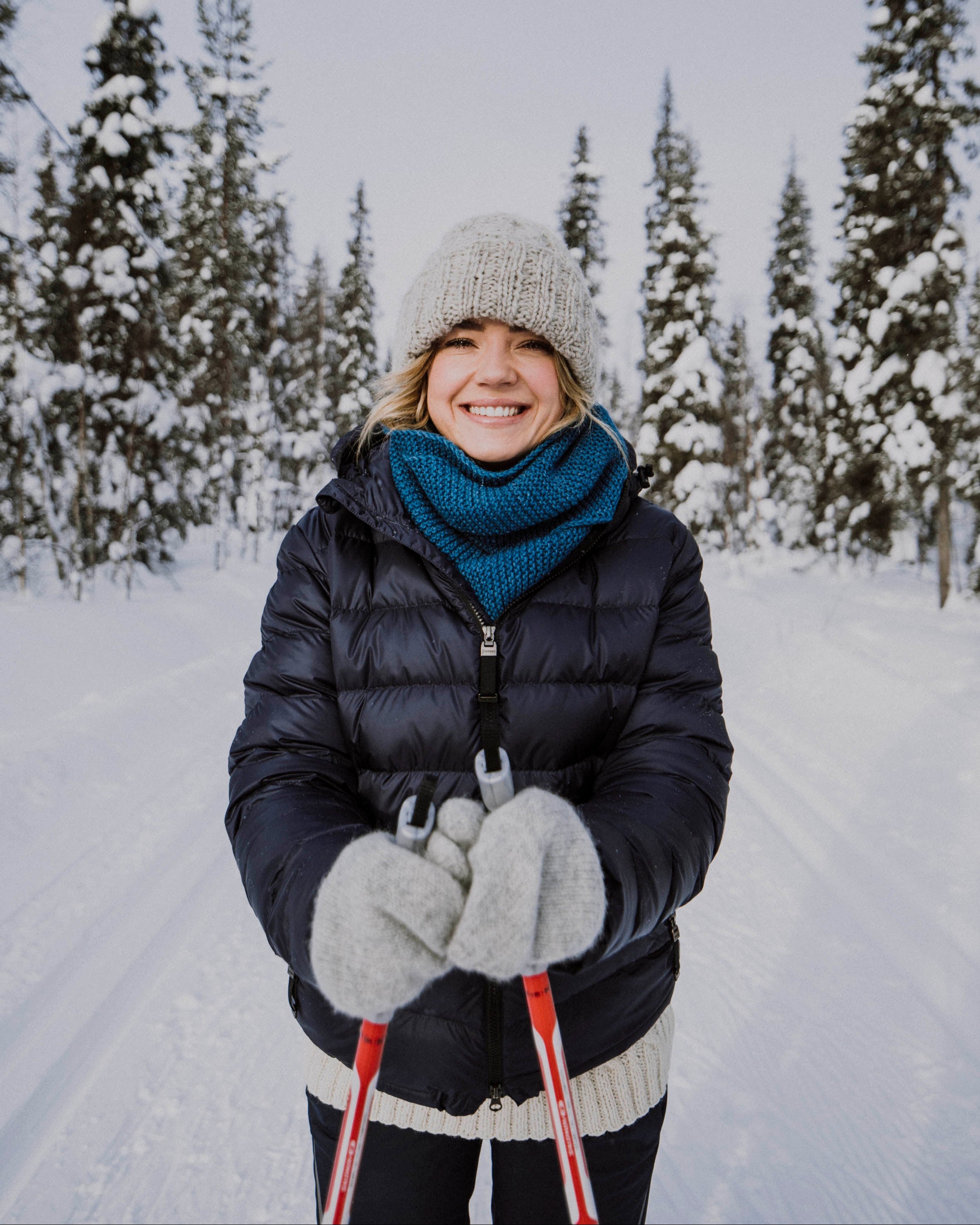 Henkilö hiihtää hymyillen lumisessa metsämaisemassa yllään tummansininen Ruska-kevytuntuvatakki (väri: pohjoisen sininen), harmaa pipo, sininen kauluri ja harmaat lapaset.