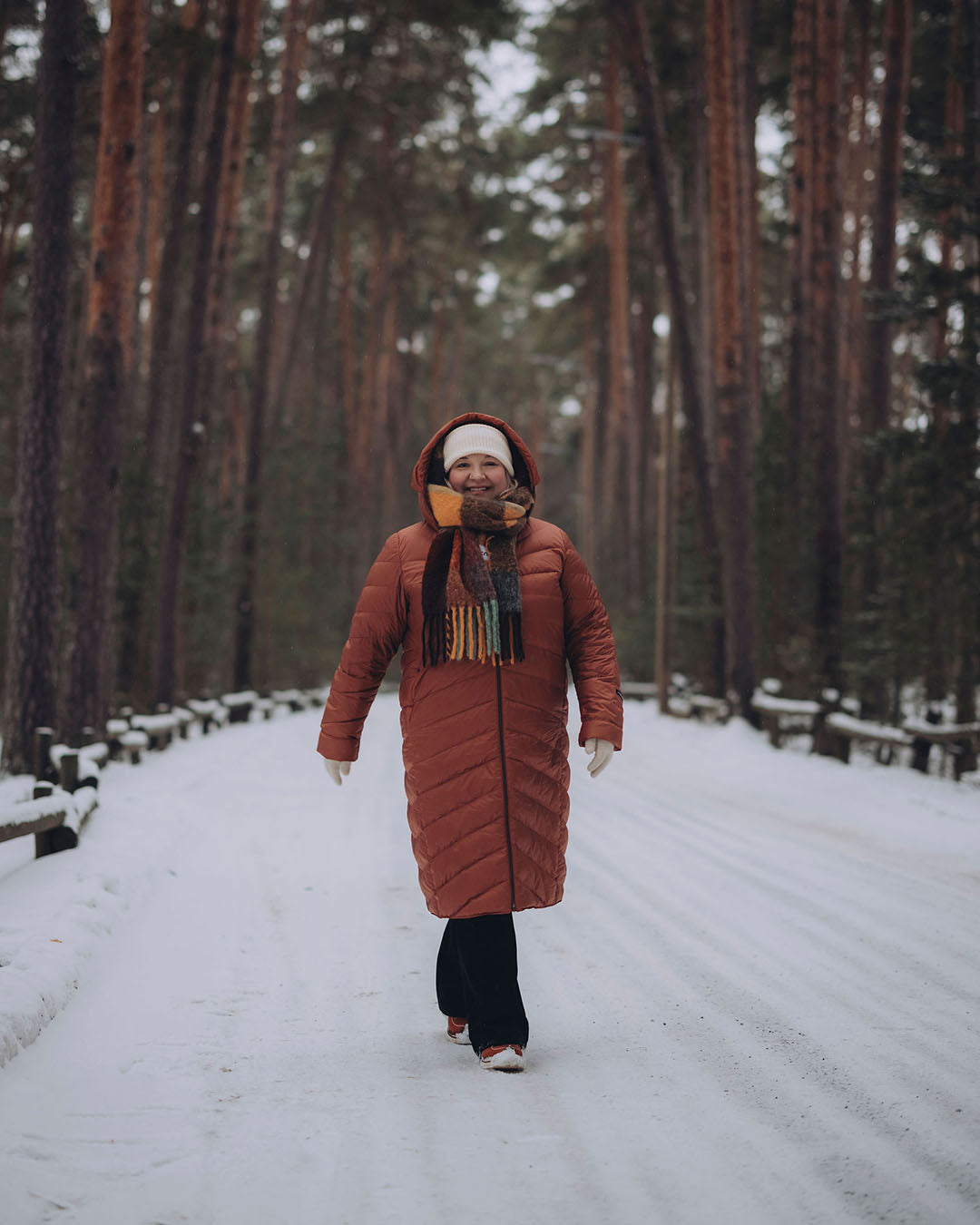 Nainen kävelee lumisella metsätiellä yllään murretun oranssi, pitkä, tikattu Säde-kevytuntuvatakki (väri: ruusukulta), vaalea pipo ja monivärinen villahuivi.