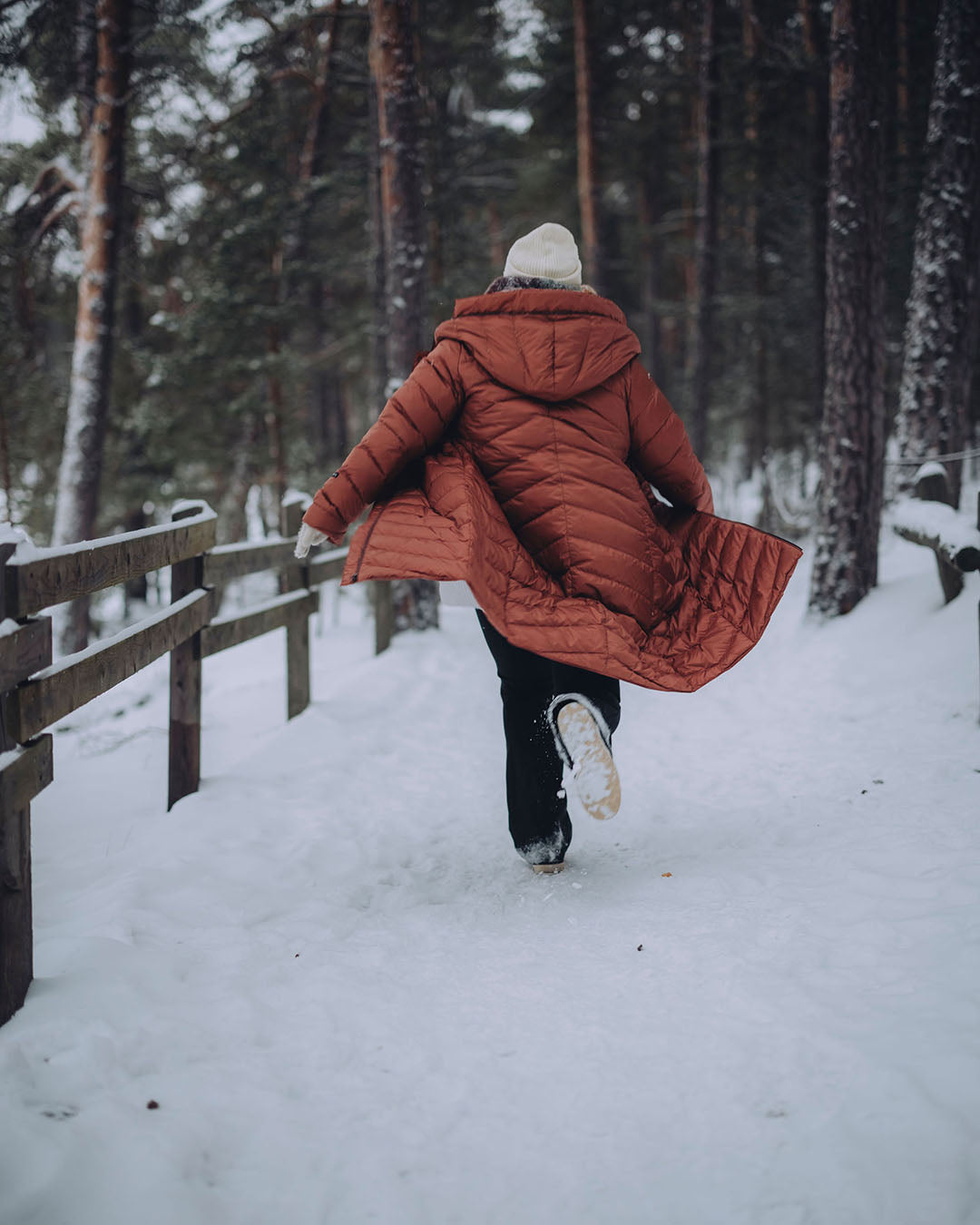 Nainen juoksee takki auki lumisella metsäpolulla murretun oranssissa, tikatussa Säde-kevytuntuvatakissa (väri: ruusukulta) ja valkoisessa pipossa. Naisen vasemmalla puolella on aita.