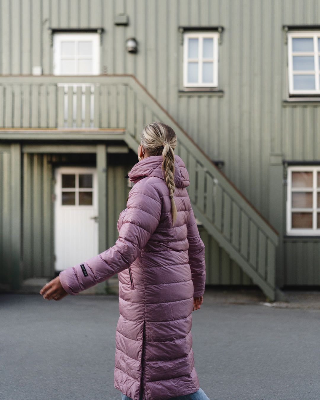Nainen kävelee ulkona selkä kameraan päin, yllään vaaleanpunainen pitkä Hanne-kevytuntuvatakki (väri: villikukka). Taustalla on vaaleanvihreä puuverhoiltu rakennus.