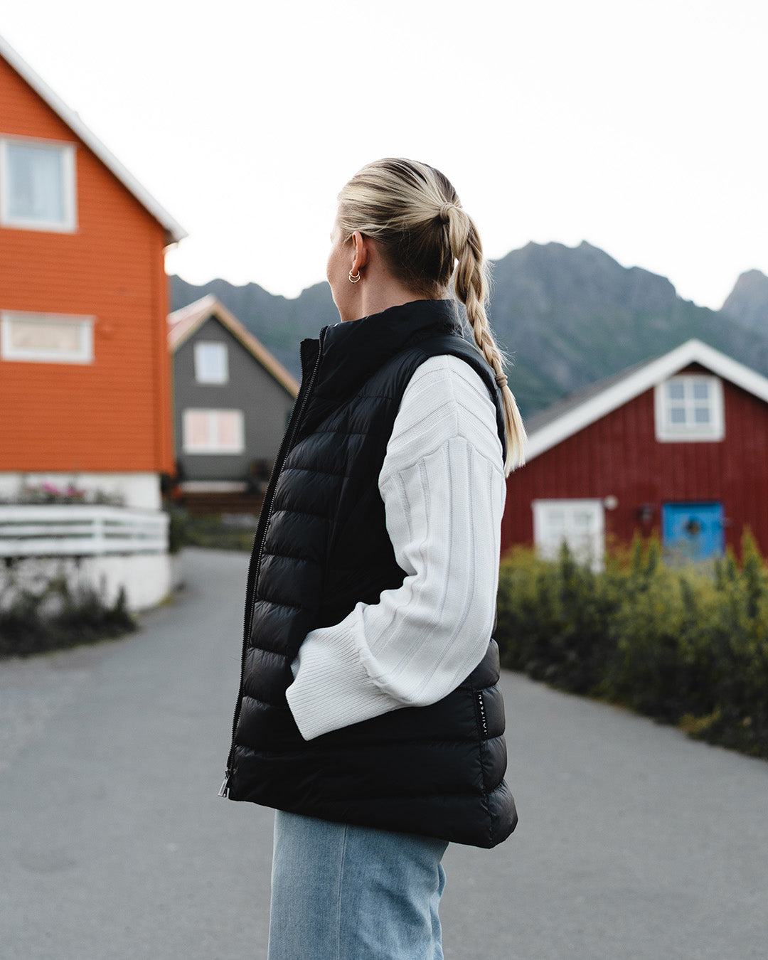 Nainen seisoo kalastajakylämaisemassa sivuttain kameraan päin. Hänellä on yllään musta Lady-untuvaliivi (väri: hiilenharmaa), valkoinen neule ja vaaleansiniset farkut.