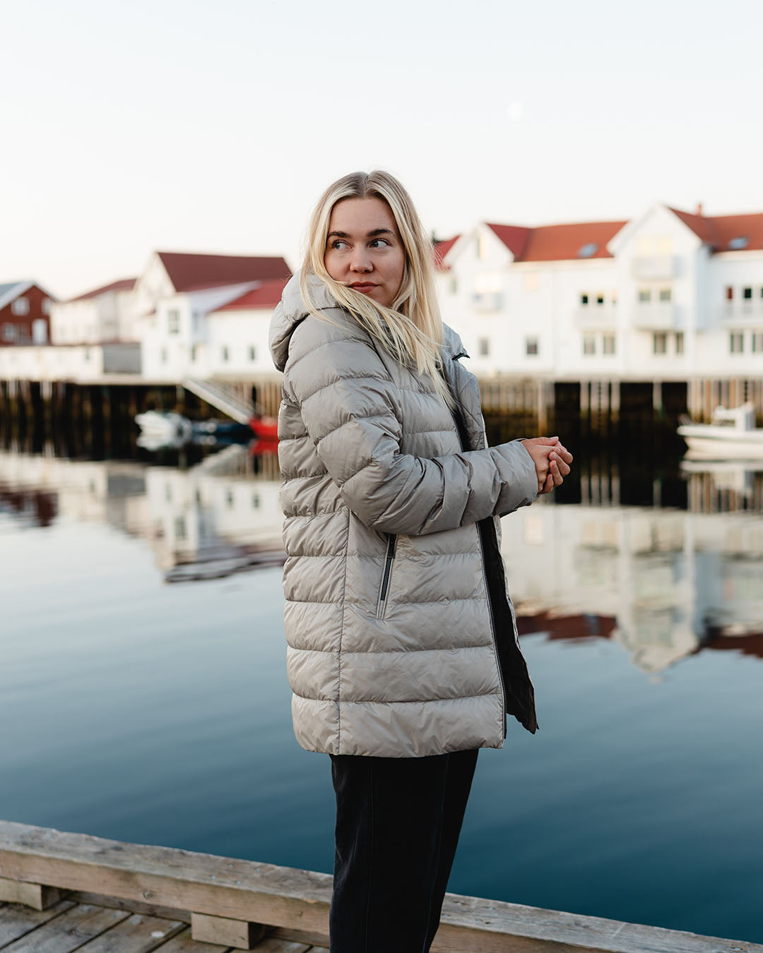 Nainen seisoo laiturilla veden äärellä taustallaan kalastajakylän vaaleita taloja ja veneitä. Hänellä on päällä beige Ruut-kevytuntuvatakki (väri: samppanja), jossa on huppu ja vetoketjulliset taskut. 
