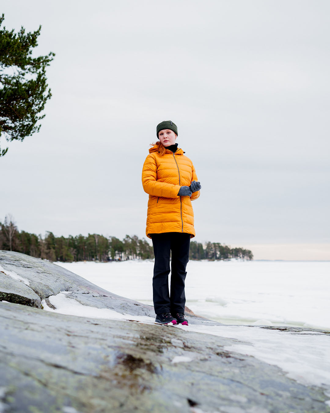 Nainen seisoo lumisella kalliolla yllään keltainen Ruut-kevytuntuvatakki (väri: meripihka), tummat housut, harmaat hansikkaat ja vihreä pipo. Taustalla näkyy jäätynyt vesistö ja metsäinen ranta.