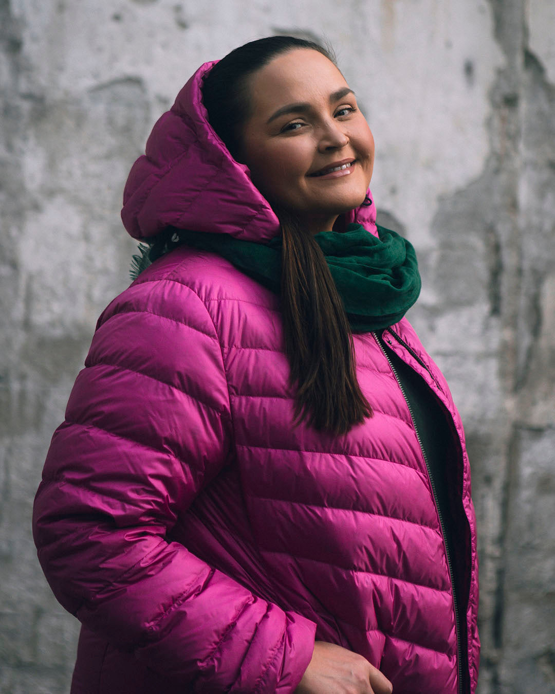 Nainen seisoo harmaan kiviseinän edessä ja hymyilee kameralle. Hänellä on yllään kirkkaan pinkki (väri: pitaija) Jasmin kevytuntuvatakki, jossa on huppu ja vinot tikkaukset. Takki on kevyesti kiiltävä, ja sen alle on puettu vihreä huivi, joka tuo lisäväriä asuun.