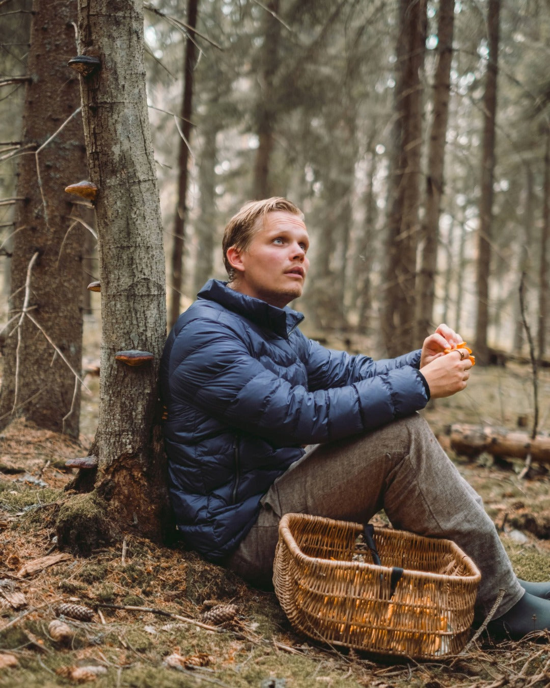 Mies istuu metsässä puuta vasten ja pitää käsissään sieniä. Hänellä on yllään sininen Hile-kevytuntuvatakki (väri: myrskyluoto) ja edessään punottu kori.
