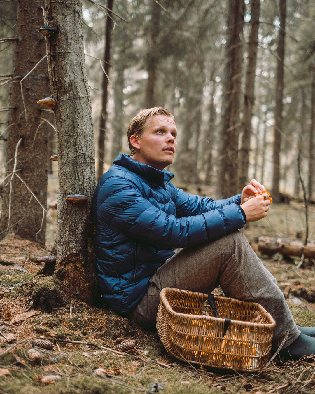 Mies istuu metsässä puuta vasten ja pitää käsissään sieniä. Hänellä on yllään sininen Hile-kevytuntuvatakki (väri: myrskyluoto) ja edessään punottu kori.