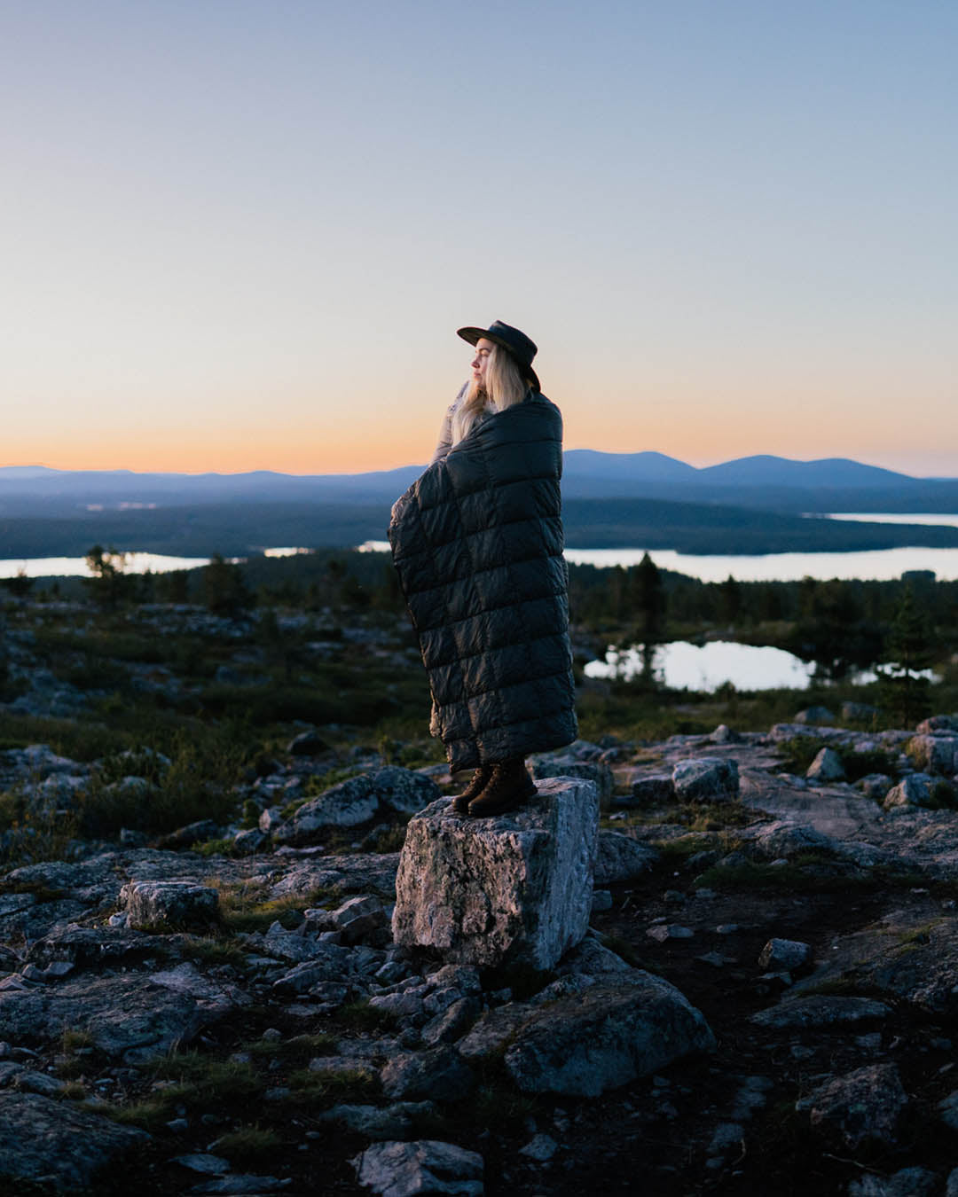 Henkilö seisoo kivellä avoimessa tunturimaisemassa auringonlaskun aikaan. Hän on kääriytynyt pitkään, tikattuun, tummanharmaaseen Kulkuri-untuvatorkkupeittoon (väri: antrasiitti) ja katsoo horisonttiin. 